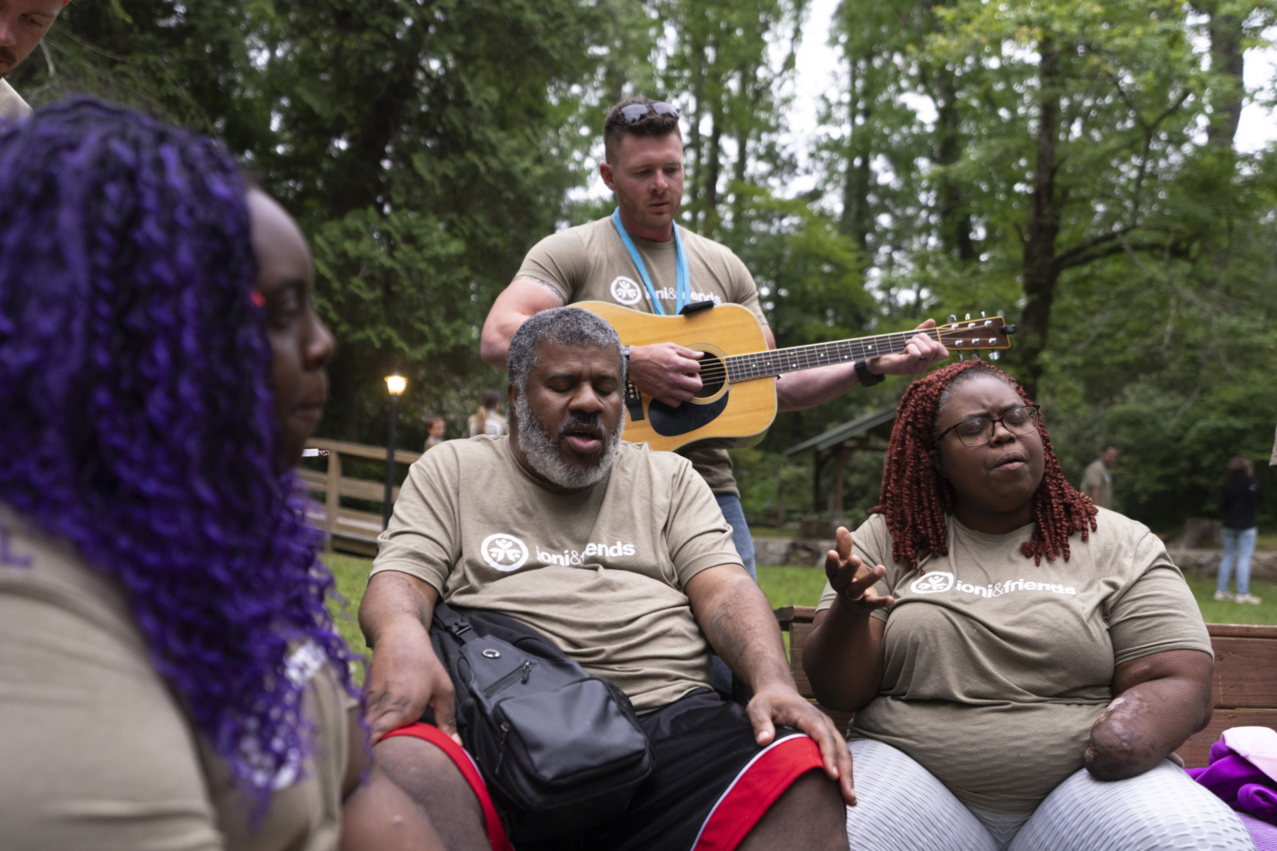 Outdoor singing circle with a man strumming a guitar while Natacha and her husband, wearing matching Joni and Friends shirts, sing along in a park setting.