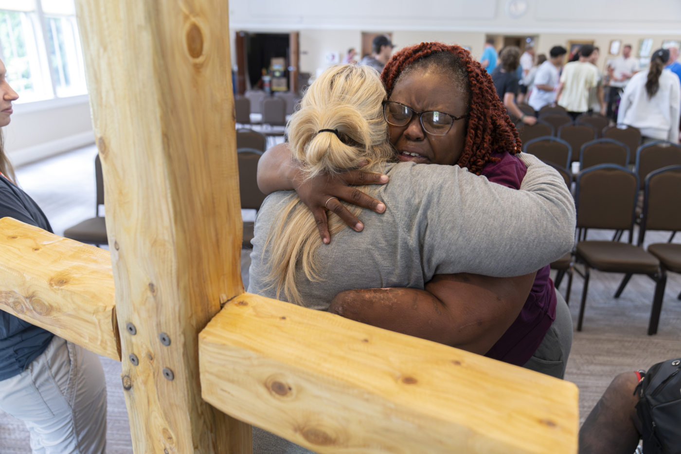 Natacha hugging a woman tightly while she is crying, with other people in the background and a wooden cross in front of them.