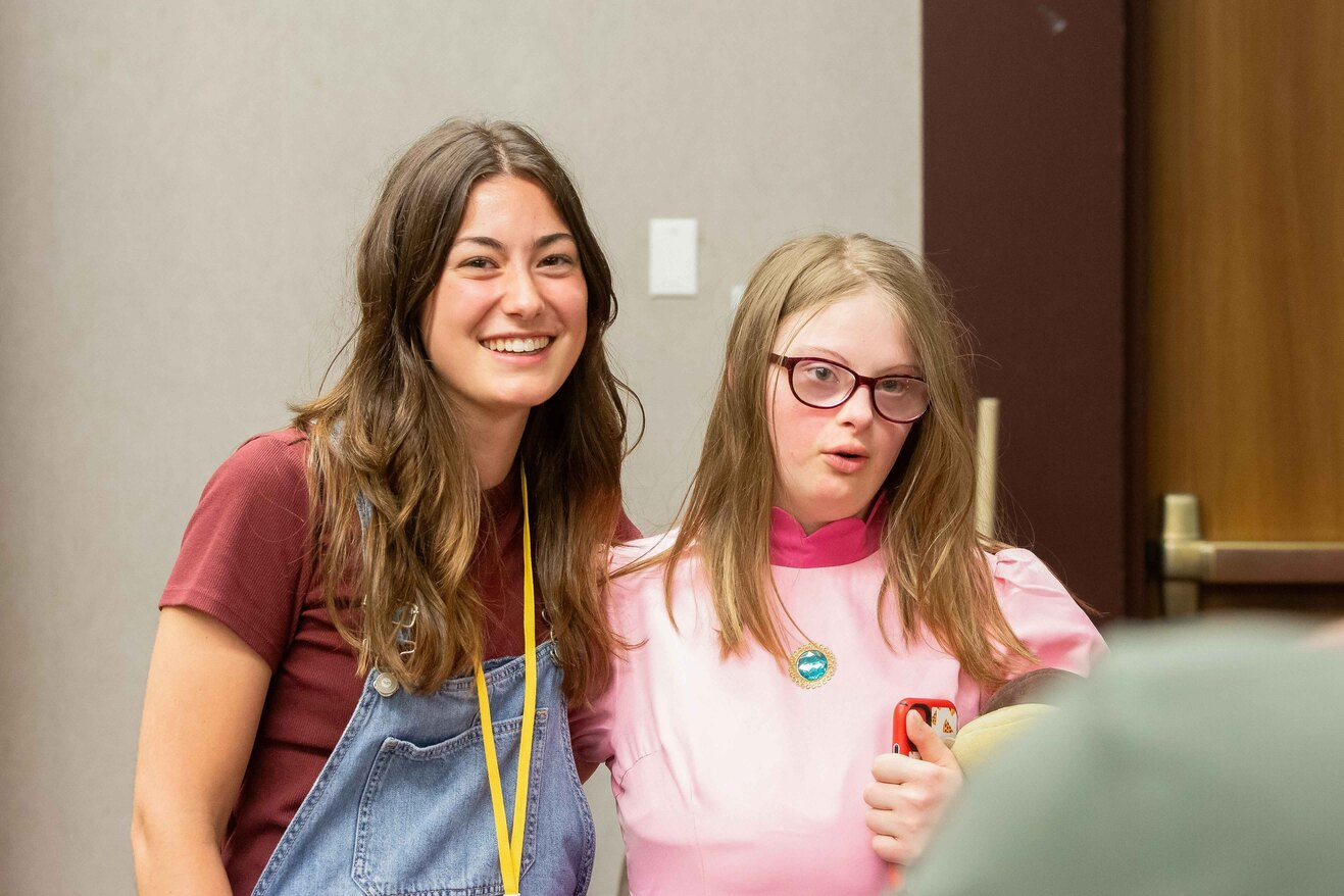 Two young women pose indoors, one with brown hair in denim overalls and a maroon shirt, the other with glasses in a pink top, smiling at the camera.