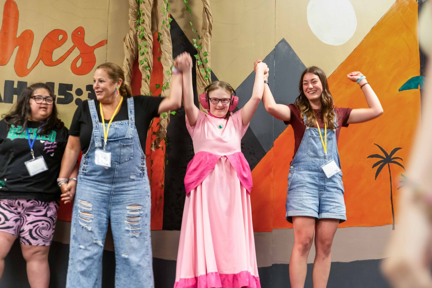 Four girls on a stage, arms raised in celebration, posing together in front of a colorful mural; center girl wears a pink dress with a darker pink sash and pink ear protectors.