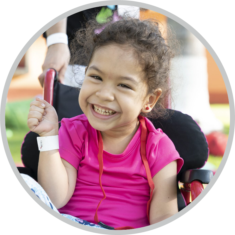 Smiling young girl in a pink shirt seated in a wheelchair outdoors, with a white wristband on her left wrist.